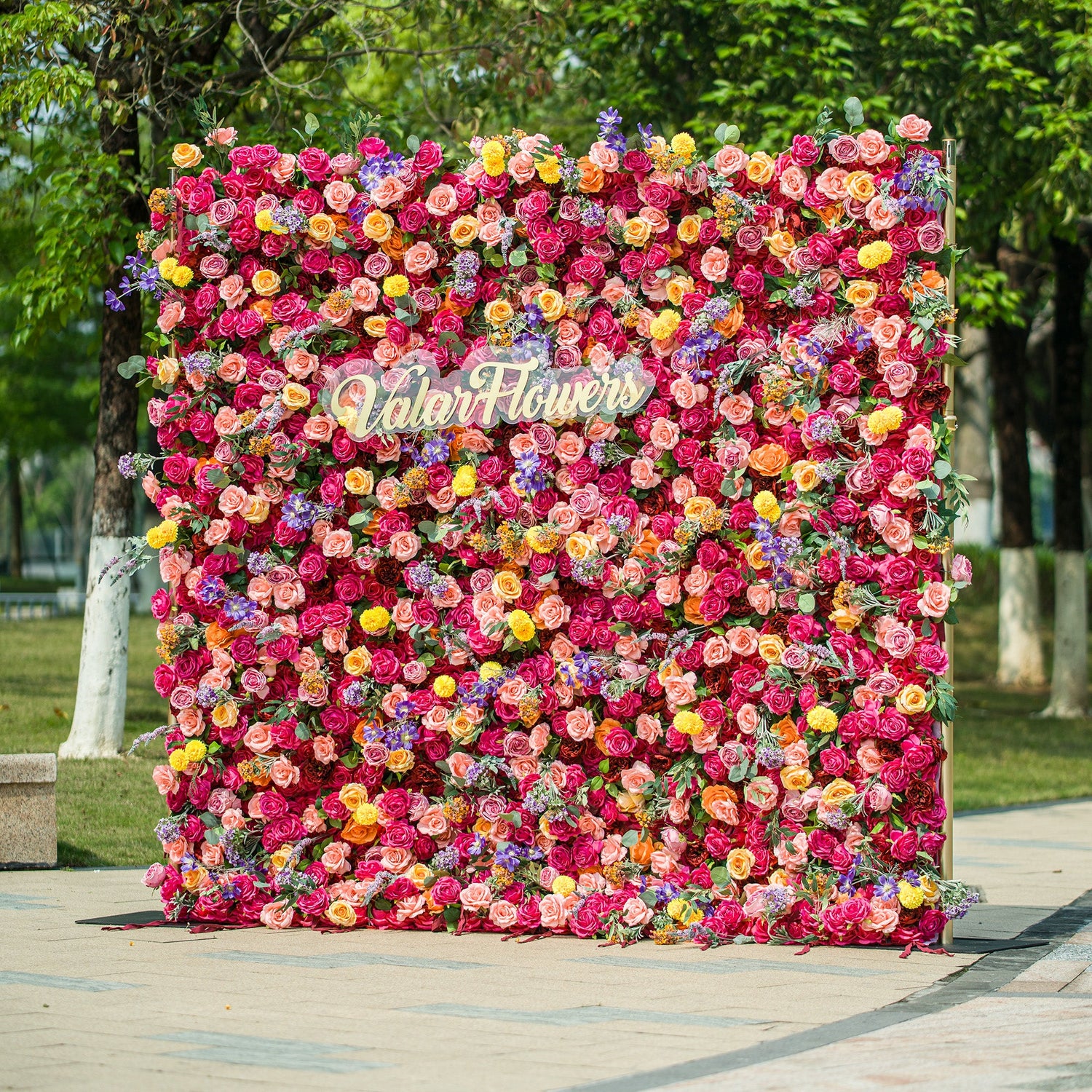 A vibrant floral wall by ValarFlowers bursts with color! Dense with roses, daisies, lavender, and more in shades of pink, red, yellow, purple—each bloom meticulously arranged. The gold “ValarFlowers” logo stands out at the center, set against lush green foliage in a park - like outdoor setting. Perfect for events, it creates a lively, Instagram - worthy backdrop full of texture and charm. 