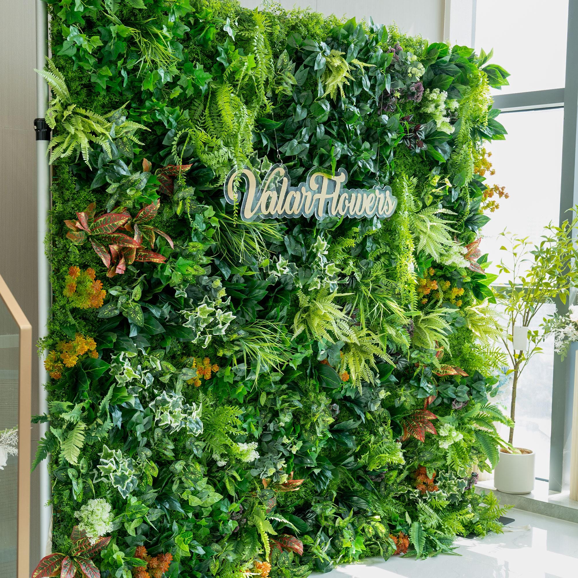This image features a lush greenery wall by ValarFlowers. The wall is a dense arrangement of various artificial plants. Prominent among them are different types of ferns, ivy - like vines, and broad - leafed foliage in varying shades of green. There are also splashes of color from orange and red - tinted plants, adding contrast. The overall effect is a vibrant, natural - looking backdrop ideal for events or interior decor. 