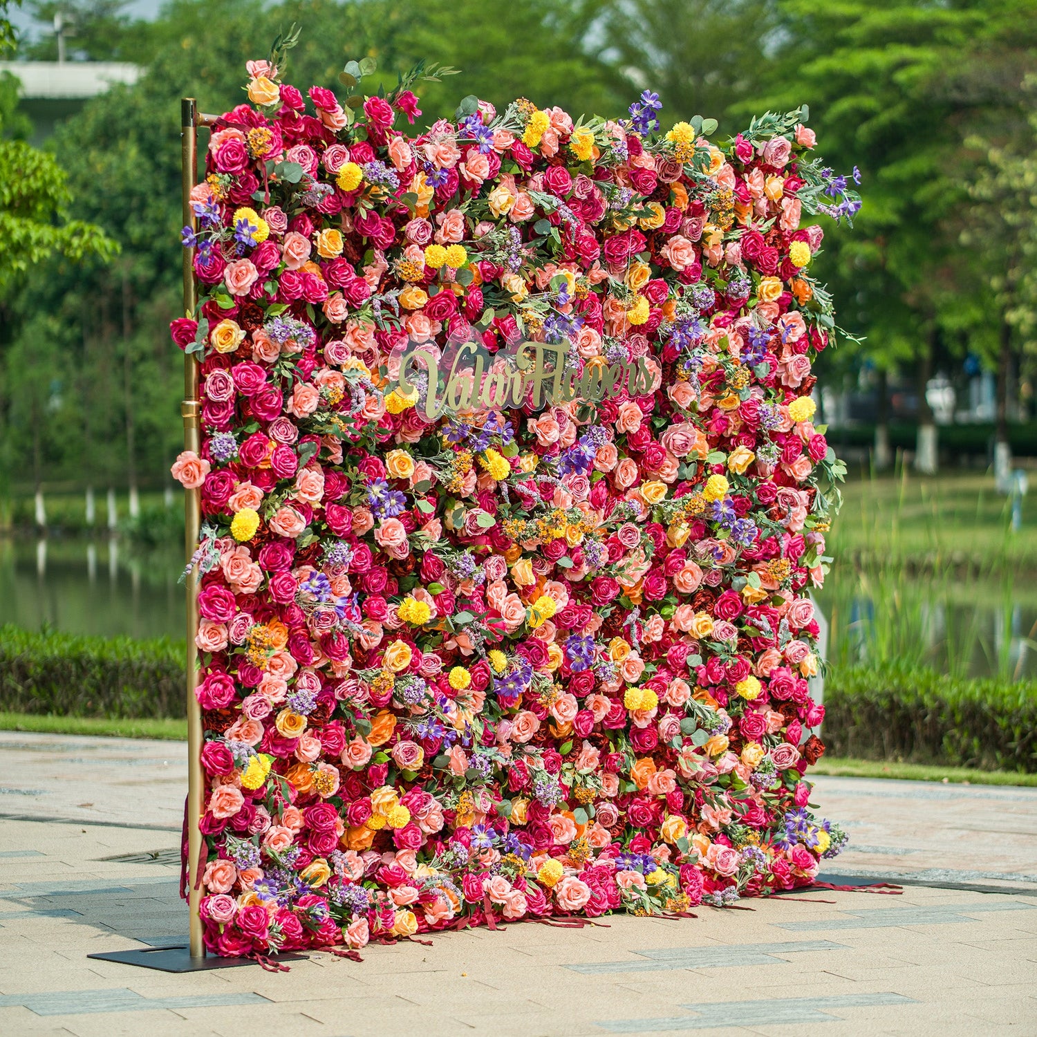A vibrant floral wall by ValarFlowers features an array of colorful blooms—roses, daisies, lavender, and more—arranged densely. The gold “ValarFlowers” logo stands out at the center. Set against a park backdrop with a lake and greenery, the wall, framed by a gold stand, bursts with hues like pink, red, yellow, and purple, creating a lively, picturesque scene perfect for events or photo ops. 