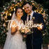This warm, intimate wedding portrait captures a smiling bride and groom posing together, holding a glowing custom neon sign that reads “The Williams”. The bride wears a lace wedding gown and veil, holding a soft pink and ivory rose bouquet, while the groom is in a navy suit with a boutonnière. They stand in front of a lush floral arch adorned with candlelight, creating a romantic, timeless atmosphere that blends modern neon decor with classic wedding elegance.