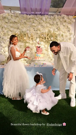 A joyful outdoor scene with a family in white - themed outfits. A young girl in a frilly dress, a woman in a voluminous gown, and a man in a white suit are in front of a white floral backdrop saying "ZAIA SKY". There's a colorful cake, suggesting a celebratory event. 