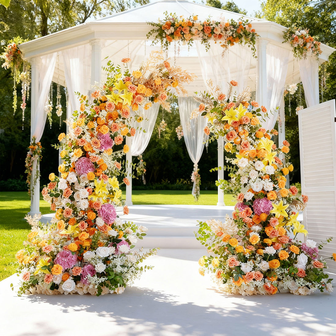 This image shows a beautifully decorated outdoor wedding gazebo. The gazebo, with white drapes, is adorned with vibrant floral arrangements in shades of orange, yellow, pink, and white. Two large, colorful floral arches frame the entrance, leading to a white platform. Set in a lush, green garden under bright sunlight, it creates a romantic and festive atmosphere, perfect for a wedding ceremony.