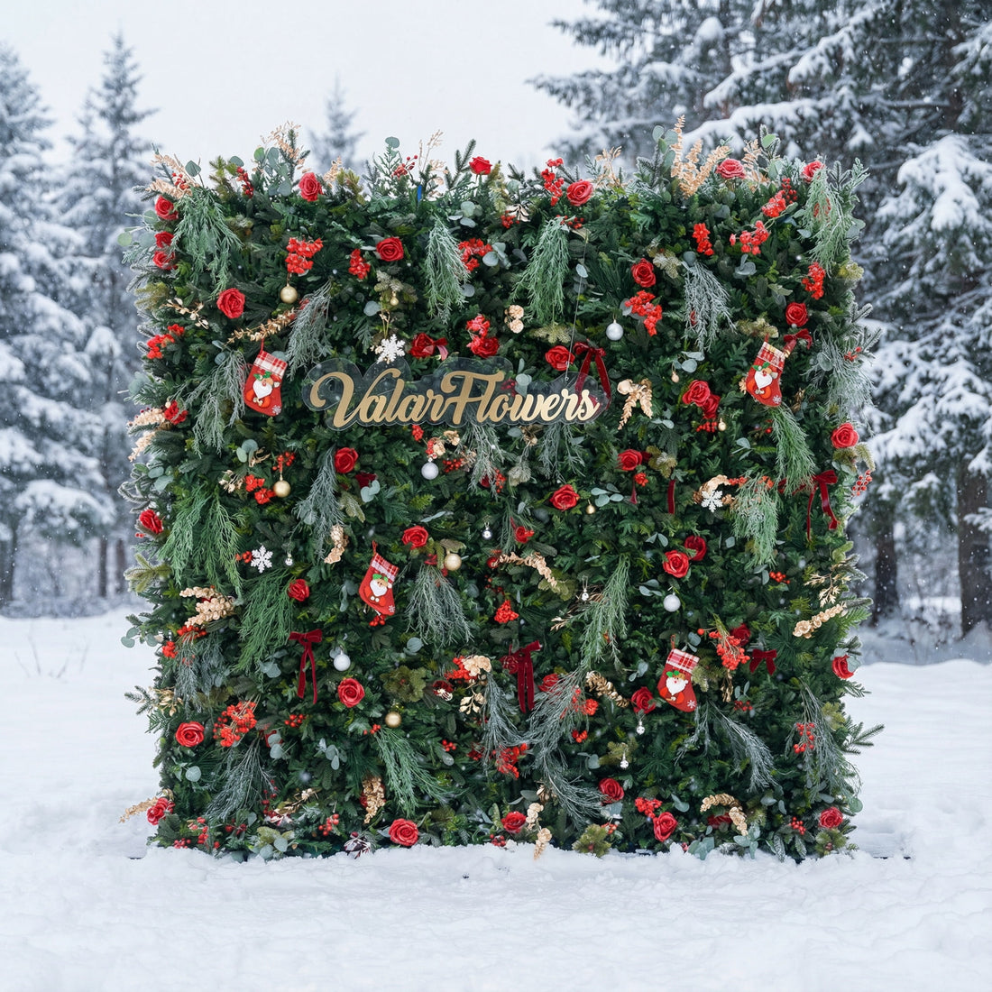 This festive square backdrop sits in a snow-covered landscape (snow-laden evergreens behind) and is dense with lush green foliage, red roses, clustered blooms, and red berries. Accents include mini Santa stockings, gold/silver ornaments, snowflakes, and a polished “ValarFlowers” sign. The rich red-and-green palette, paired with wintry snow, evokes cozy holiday warmth—ideal for Christmas events, festive photo ops, or seasonal outdoor decor.