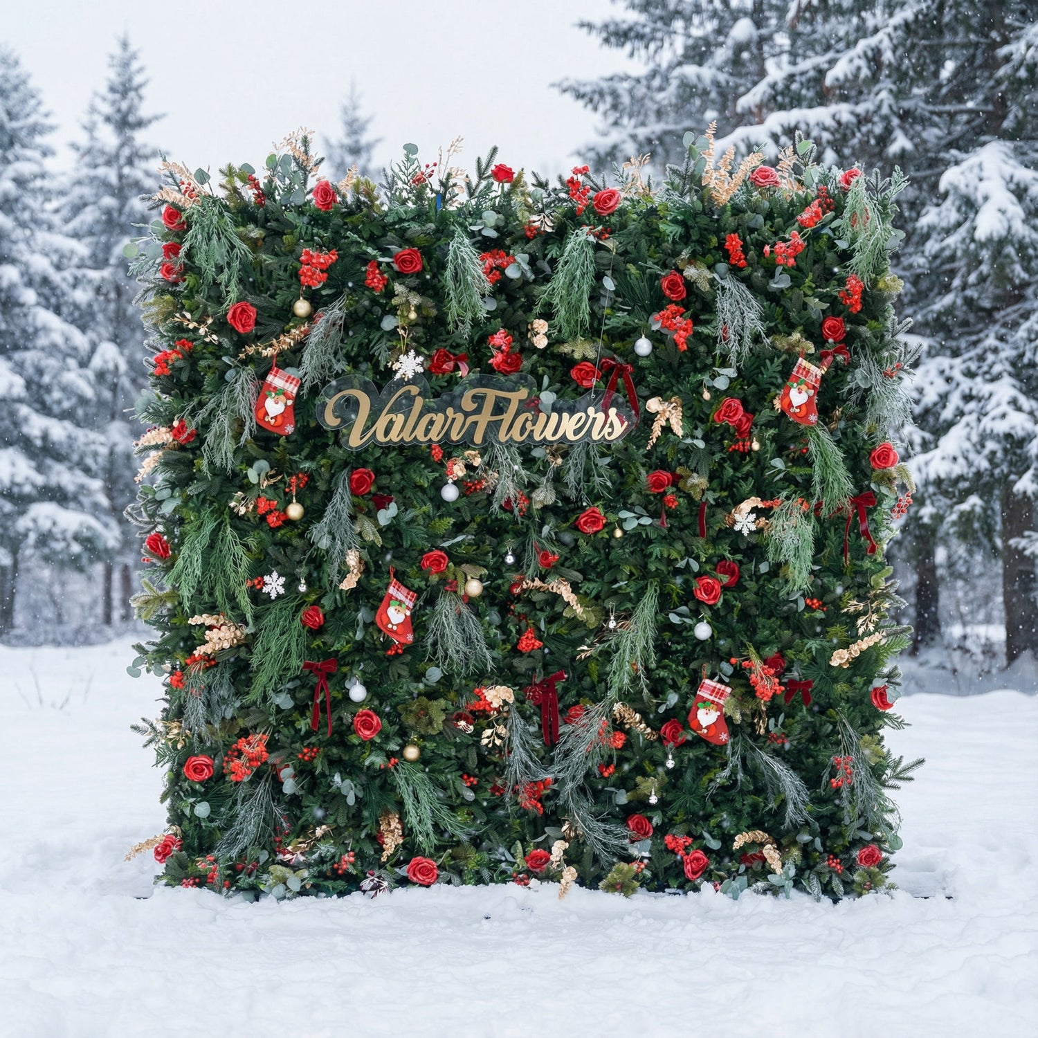 This festive square backdrop sits in a snow-covered landscape (snow-laden evergreens behind) and is dense with lush green foliage, red roses, clustered blooms, and red berries. Accents include mini Santa stockings, gold/silver ornaments, snowflakes, and a polished “ValarFlowers” sign. The rich red-and-green palette, paired with wintry snow, evokes cozy holiday warmth—ideal for Christmas events, festive photo ops, or seasonal outdoor decor.