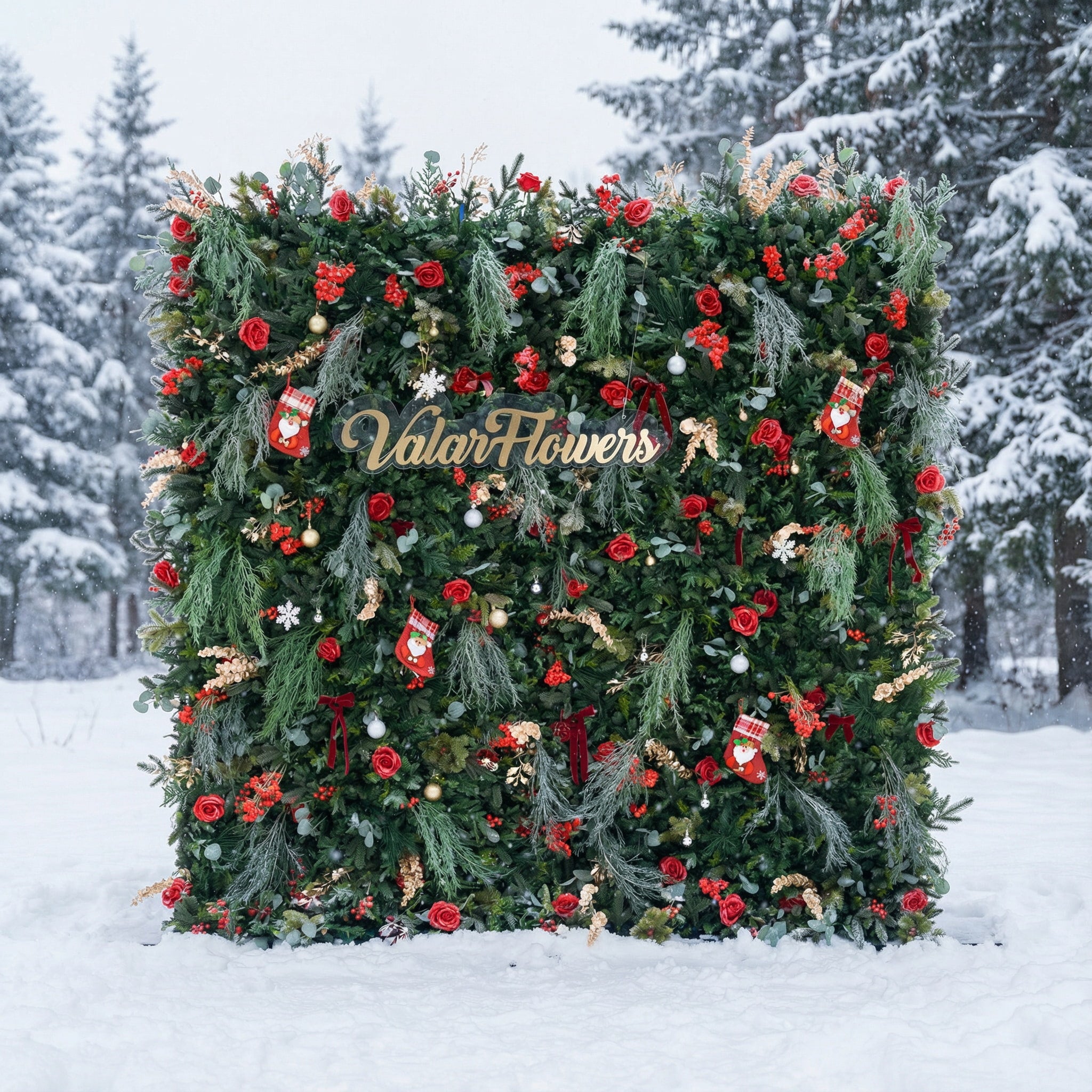 This festive square backdrop sits in a snow-covered landscape (snow-laden evergreens behind) and is dense with lush green foliage, red roses, clustered blooms, and red berries. Accents include mini Santa stockings, gold/silver ornaments, snowflakes, and a polished “ValarFlowers” sign. The rich red-and-green palette, paired with wintry snow, evokes cozy holiday warmth—ideal for Christmas events, festive photo ops, or seasonal outdoor decor.
