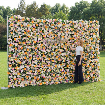This large floral backdrop (set on sunlit grass, tree-lined background) bursts with soft blooms: pale pink, white, and warm orange daisy-like flowers, clustered lushly. A polished "ValarFlowers" sign anchors the design, while a woman (white top, black wide-leg pants) adjusts it, adding a dynamic touch. The pastel-and-orange palette, paired with natural greenery, evokes a cheerful, vibrant vibe—ideal for outdoor events, wedding decor, or festive photo backdrops.