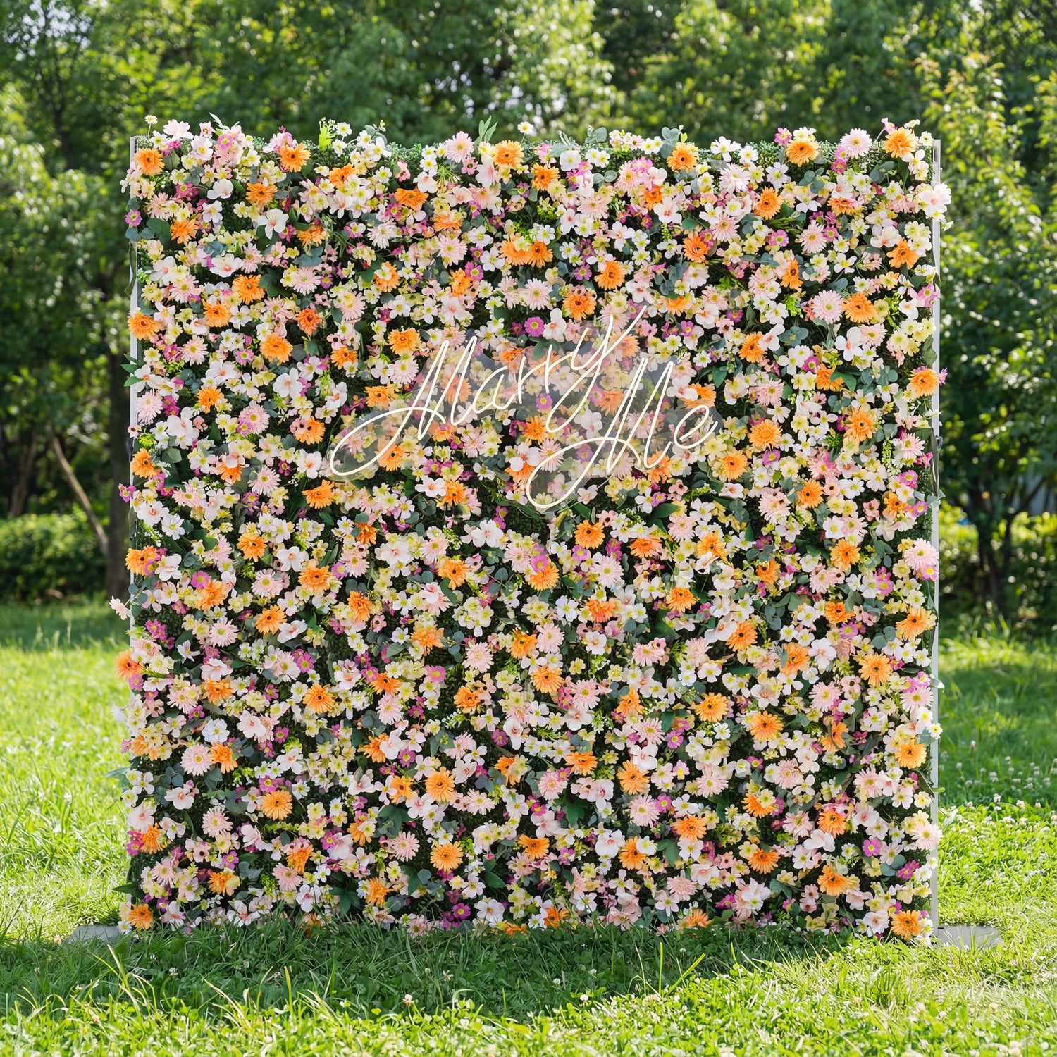 This square floral backdrop sits on sunlit grass (tree-filled background) and is dense with soft blooms: pale pink, white, and warm orange daisy-like flowers, clustered lushly. A glowing "Marry Me" sign (cursive, light-lit) anchors the center, adding romantic charm. The pastel-and-orange palette, paired with natural greenery, evokes a cheerful, dreamy vibe—ideal for proposal setups, outdoor weddings, or romantic event decor.