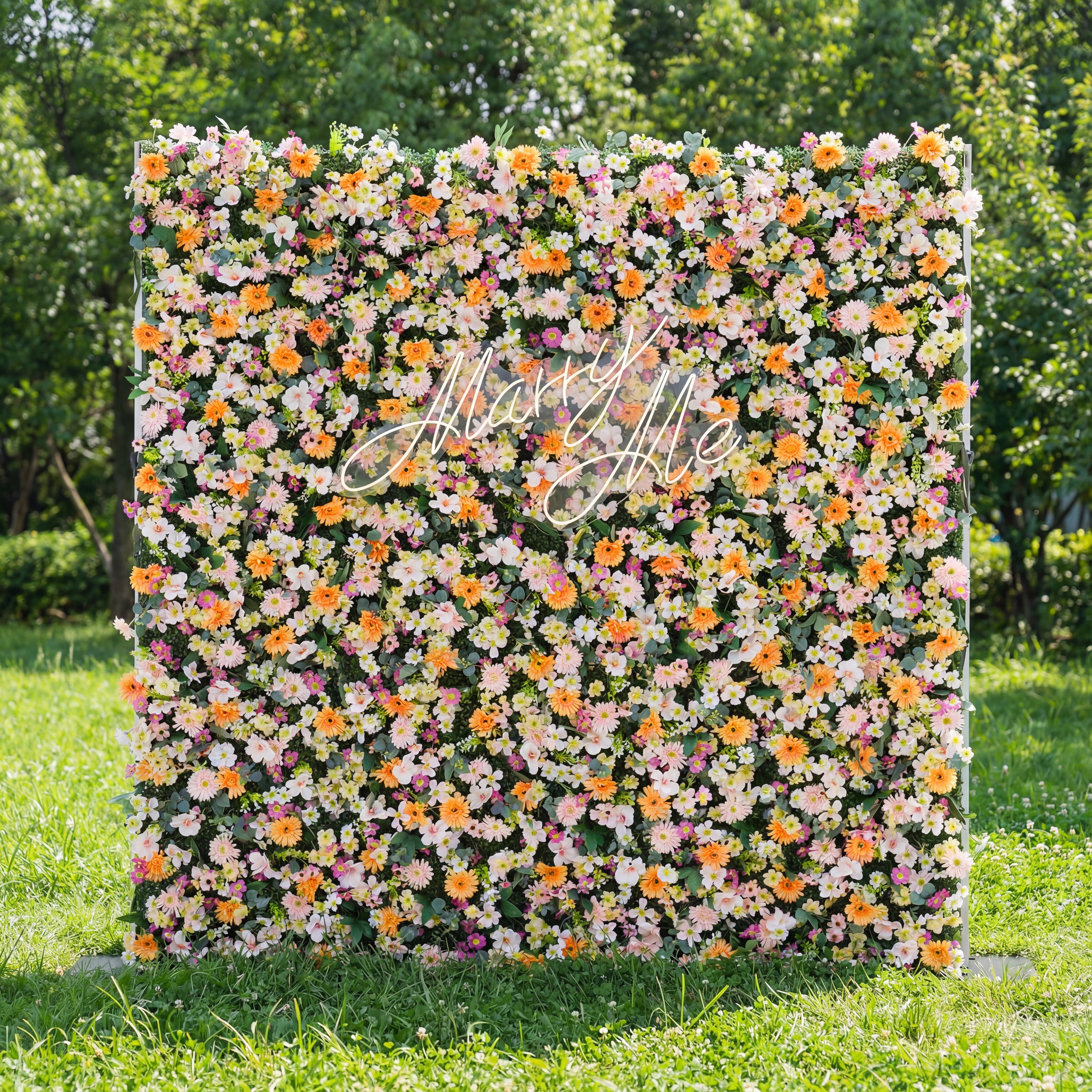 This square floral backdrop sits on sunlit grass (tree-filled background) and is dense with soft blooms: pale pink, white, and warm orange daisy-like flowers, clustered lushly. A glowing "Marry Me" sign (cursive, light-lit) anchors the center, adding romantic charm. The pastel-and-orange palette, paired with natural greenery, evokes a cheerful, dreamy vibe—ideal for proposal setups, outdoor weddings, or romantic event decor.