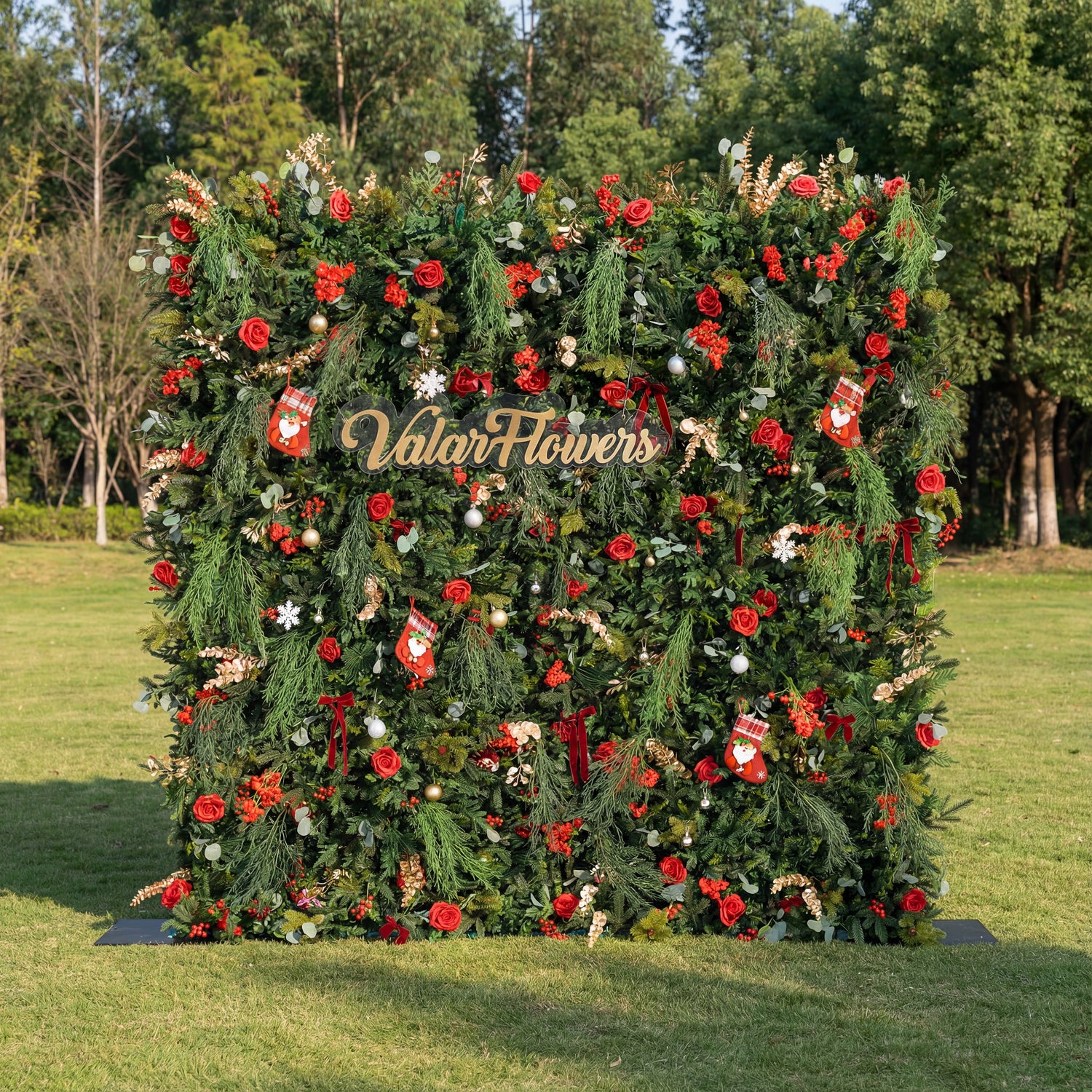 This square festive backdrop (set on sunlit grass, tree-filled background) is dense with lush green foliage, red roses, clustered blooms, and red berries. Accents include mini Santa stockings, gold/silver ornaments, snowflakes, and a polished “ValarFlowers” sign. The rich red-and-green palette, layered textures, and natural sunlight evoke cozy holiday charm—ideal for Christmas events, festive photo ops, or seasonal outdoor decor.