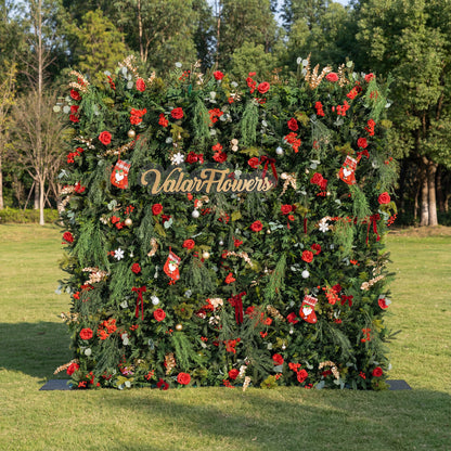 This square festive backdrop (set on sunlit grass, tree-filled background) is dense with lush green foliage, red roses, clustered blooms, and red berries. Accents include mini Santa stockings, gold/silver ornaments, snowflakes, and a polished “ValarFlowers” sign. The rich red-and-green palette, layered textures, and natural sunlight evoke cozy holiday charm—ideal for Christmas events, festive photo ops, or seasonal outdoor decor.