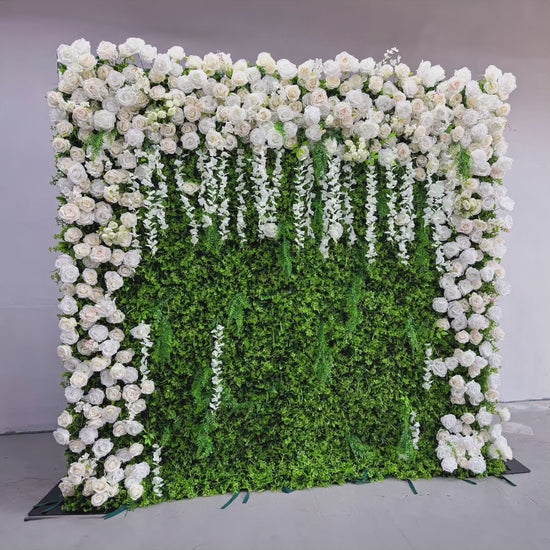 The flower wall features a green foliage base with numerous white roses. The roses are arranged to cover the top and sides, while white floral strands hang down the front. The combination of green and white creates a fresh and elegant look.
