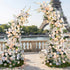 A romantic, asymmetrical floral arch from Valar Flowers, crafted with a soft, warm palette of blush pink, peach, and creamy white blooms—including roses, peonies, and delicate foliage—set against a Parisian riverside terrace with the iconic Eiffel Tower in the background. Scattered rose petals and elegant white chairs enhance the dreamy, sophisticated atmosphere, perfect for luxury weddings and editorial photoshoots.