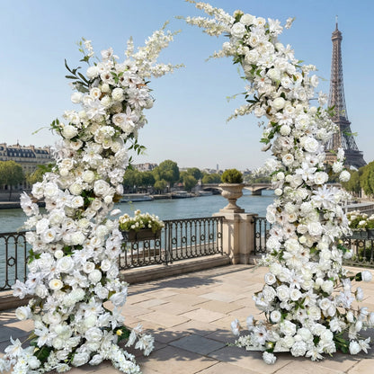 A timeless, symmetrical floral arch from Valar Flowers, crafted with a pure white palette of lush roses, lilies, and delicate foliage. Set on a sunlit Parisian riverside terrace with the iconic Eiffel Tower in the background, it creates a serene, elegant, and ethereal aesthetic ideal for luxury weddings and refined photoshoots.