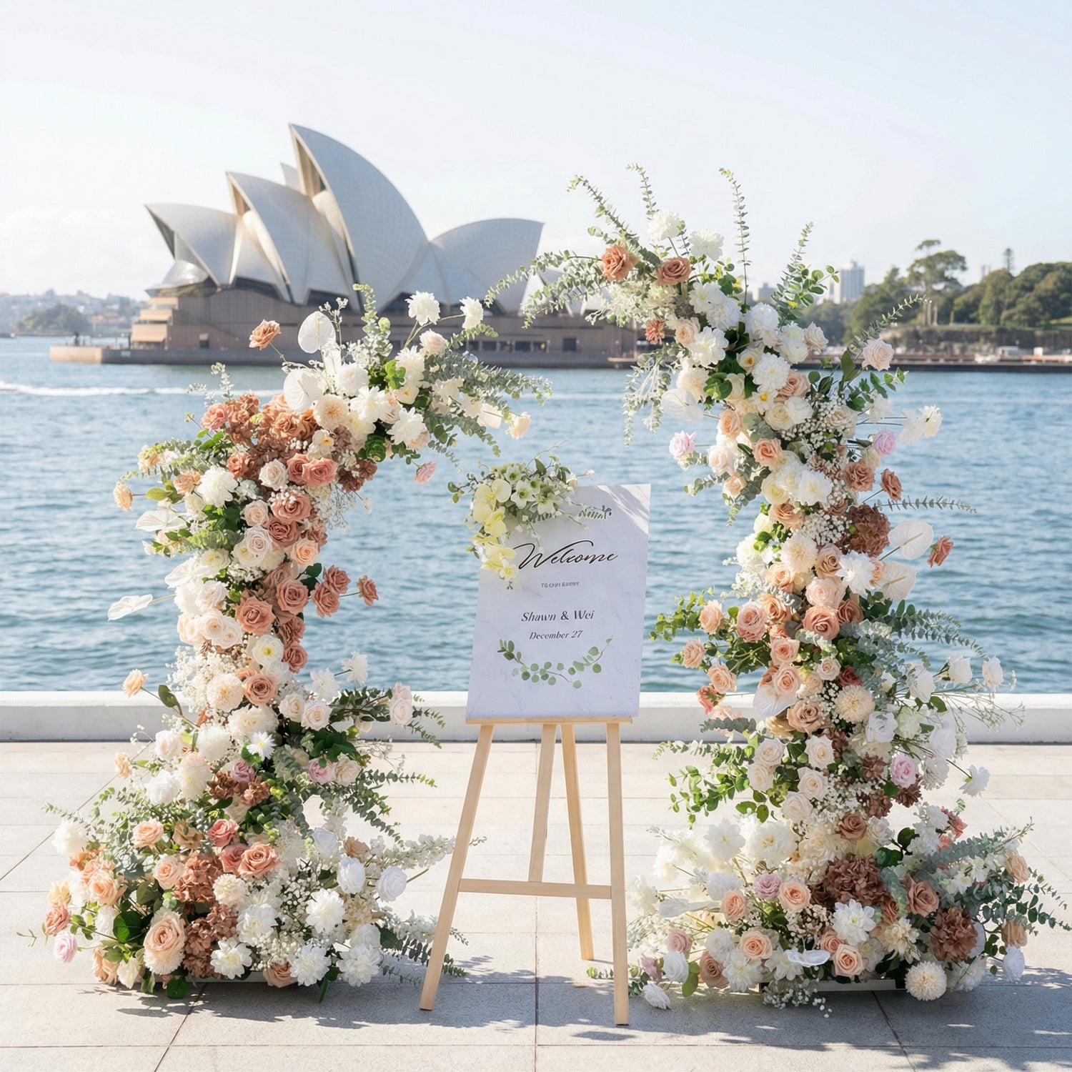 A romantic asymmetrical floral arch from **Valar Flowers**, crafted with a soft palette of blush pink, terracotta, and creamy white blooms—including roses, peonies, and delicate foliage—set against the iconic Sydney Opera House and glistening harbor. A "Welcome" sign on a wooden easlerests between the arch, creating an elegant, dreamy atmosphere perfect for coastal weddings and refined celebrations.