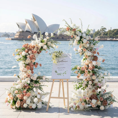 A romantic asymmetrical floral arch from **Valar Flowers**, crafted with a soft palette of blush pink, terracotta, and creamy white blooms—including roses, peonies, and delicate foliage—set against the iconic Sydney Opera House and glistening harbor. A "Welcome" sign on a wooden easlerests between the arch, creating an elegant, dreamy atmosphere perfect for coastal weddings and refined celebrations.