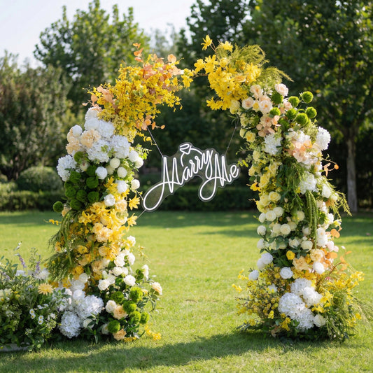 This vibrant outdoor floral arch bursts with a sunlit palette of golden yellow, crisp white, and fresh green blooms, including hydrangeas, roses, and delicate filler flowers. At its center, a glowing "Marry Me" neon sign takes the spotlight. Set on a lush green lawn framed by leafy trees, the arrangement exudes joyful romance, making it the perfect focal point for a heartfelt proposal, engagement photoshoot, or sunny celebration.