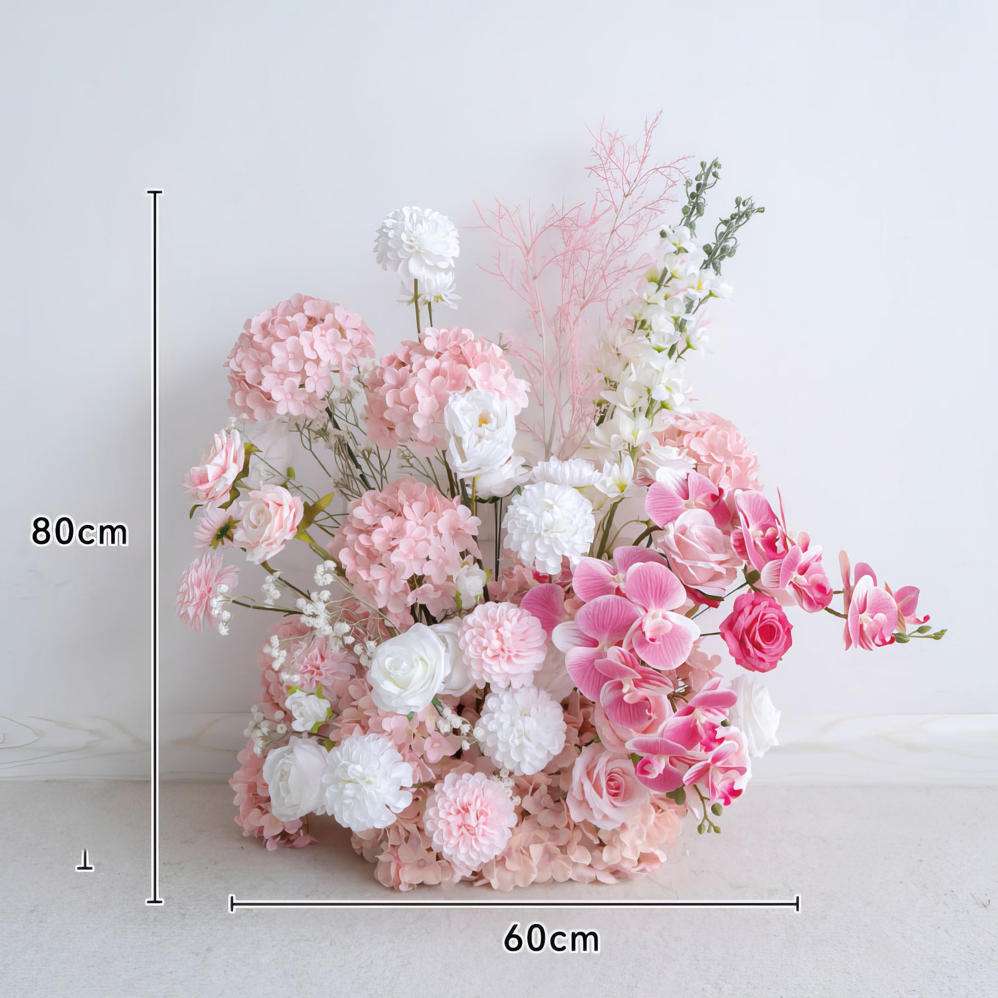 This 80cm tall, 60cm wide floral arrangement features a romantic blend of soft pink hydrangeas, blush roses, pink orchids, and crisp white blooms, accented with delicate filler flowers and airy pink foliage. The lush, layered design creates a dreamy, three-dimensional texture, making it a stunning focal point for weddings, engagement photoshoots, or romantic celebrations against a minimalist white backdrop.
