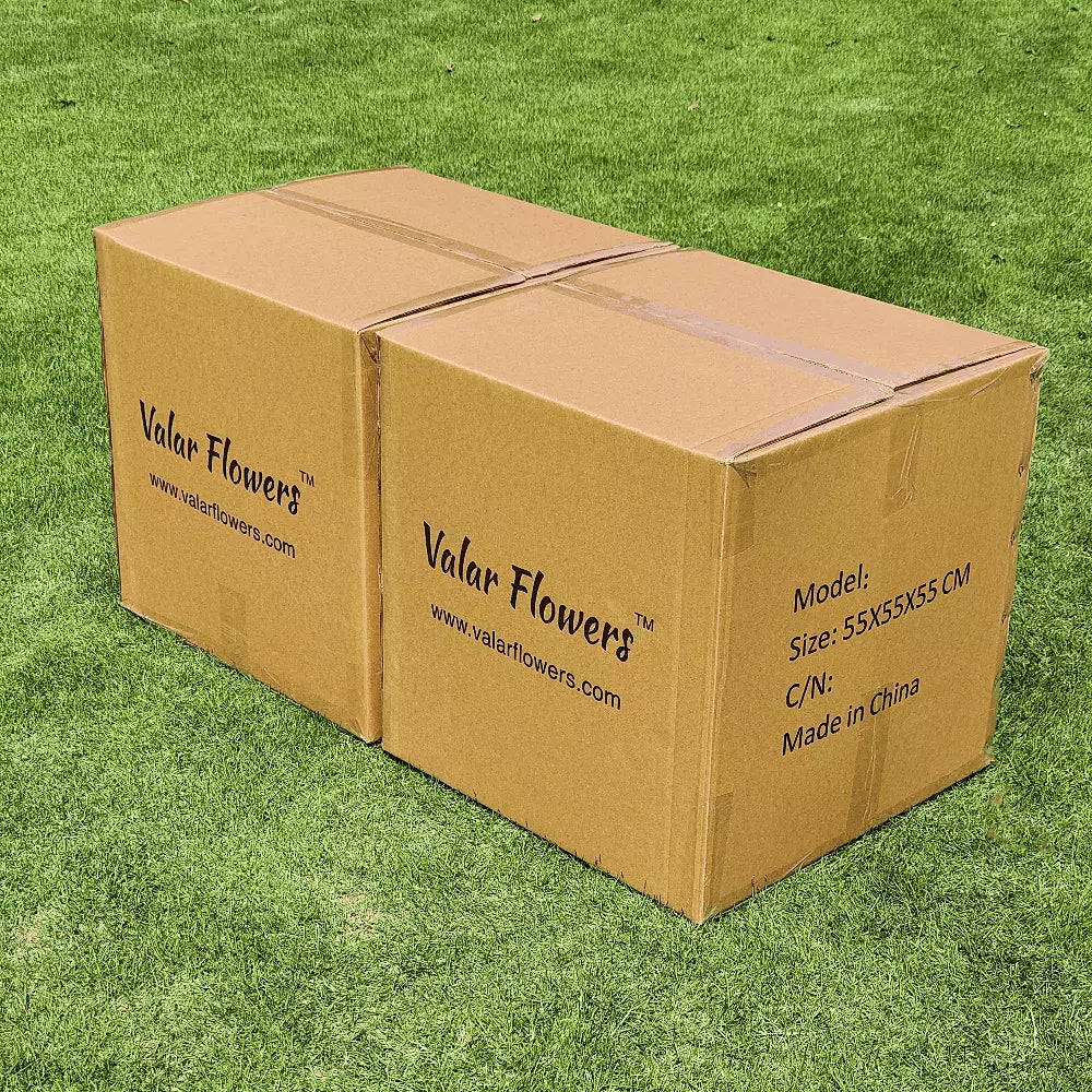 This photo shows two brown cardboard boxes labeled "Valar Flowers" with the website "www.valarflowers.com", model details "Size: 55X55X55 CM", and "Made in China". Set on green grass, it's related to floral products, likely for shipping or storage. 