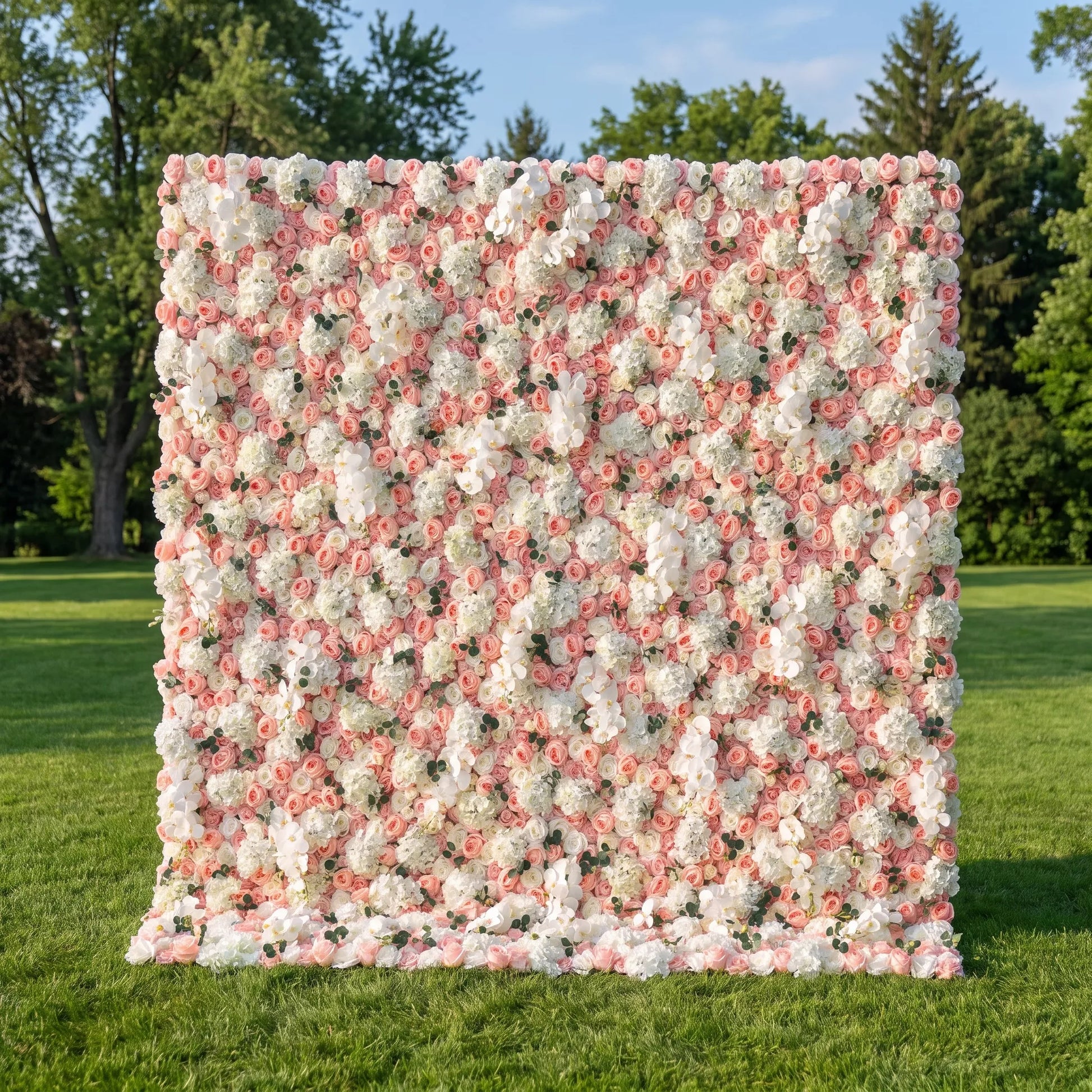 Lush full-size artificial flower wall layered with soft blush roses, large white hydrangeas and orchid blooms, placed on a sunlit green lawn with leafy trees behind. Dense, uniform coverage creates gentle romantic texture. This reusable, low-maintenance floral backdrop suits wedding photo booths, bridal showers, baby showers and pastel-themed content shoots, offering soft pink-and-white tones for elegant, Instagram-ready event decor.