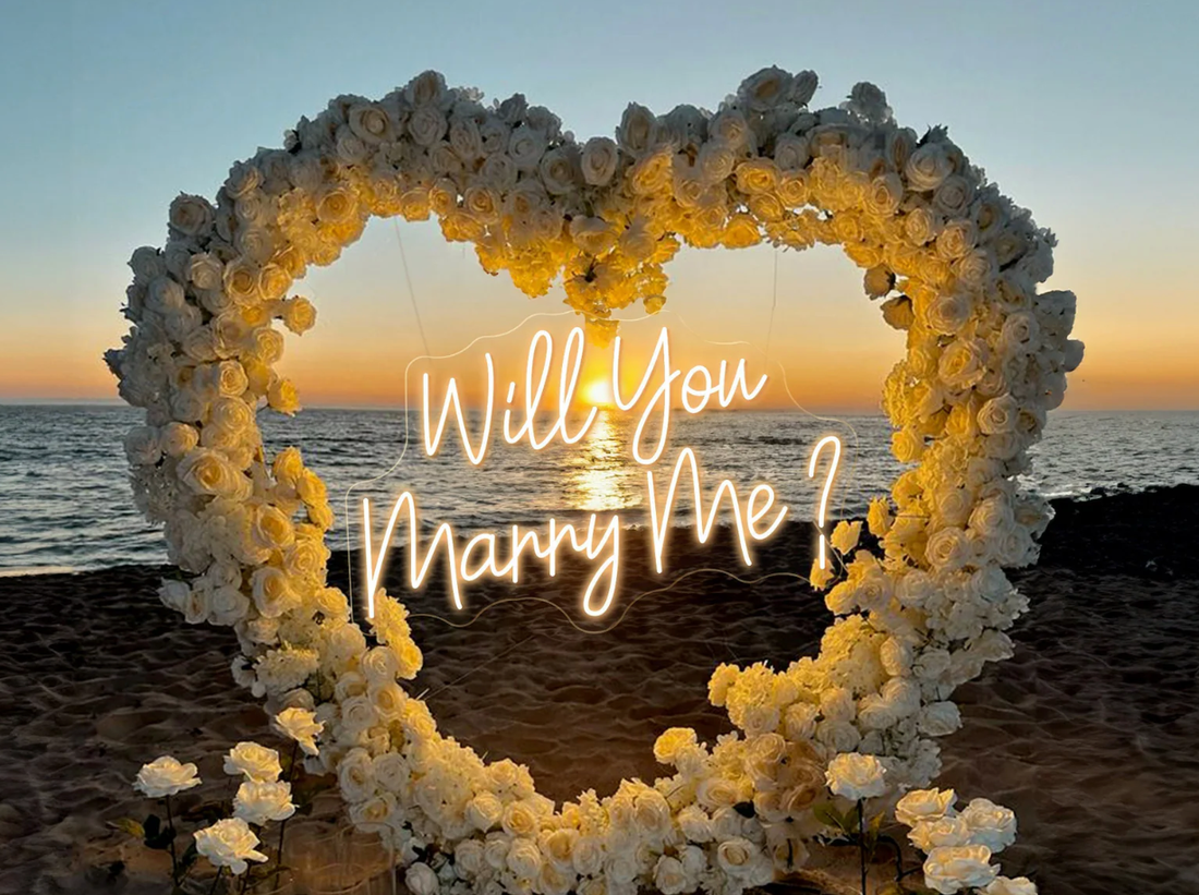 This photo from ValarFlowers captures a romantic beach proposal setup at sunset. A heart - shaped structure is densely adorned with white roses, creating a dreamy and elegant frame. Inside the heart, a glowing neon sign reads “Will You Marry Me?”. The serene ocean and the warm hues of the setting sun in the background add to the magical atmosphere. Ideal for a “Sunset Serenade” - themed proposal, it exudes love and romance.
